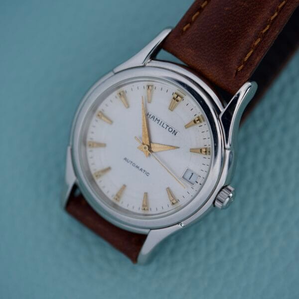 A Hamilton Automatic Date Ref. 6239 watch, featuring 25 jewels, a stainless steel case, brown leather strap, is displayed on a light blue background.
