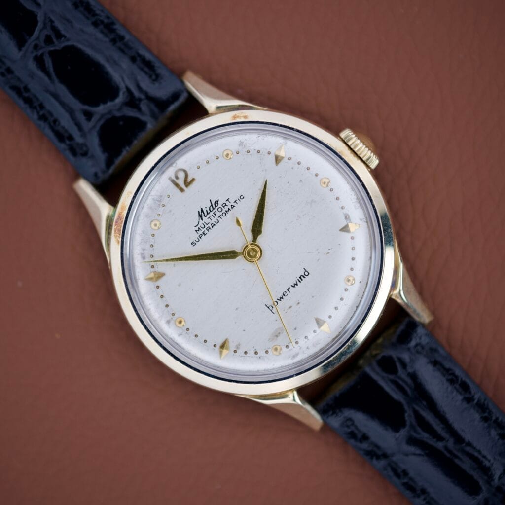 Mido Multifort Super Automatic Powerwind 14k Gold Capped Cal. 917R wristwatch with a white face and black leather strap, shown on a brown background.
