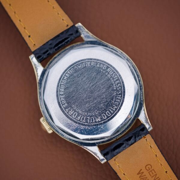 Close-up of a scratched Mido Multifort Super Automatic Powerwind 14k Gold Capped Cal. 917R watch back with engraved text, resting on a brown leather surface.