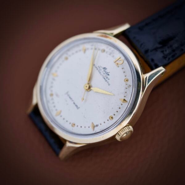 A Mido Multifort Super Automatic Powerwind 14k Gold Capped Cal. 917R with a white dial and black leather strap is displayed on a brown surface.
