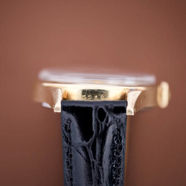 Close-up of a Mido Multifort Super Automatic Powerwind 14k Gold Capped Cal. 917R watch case, showing the number 5244 above a black leather strap on a brown background.
