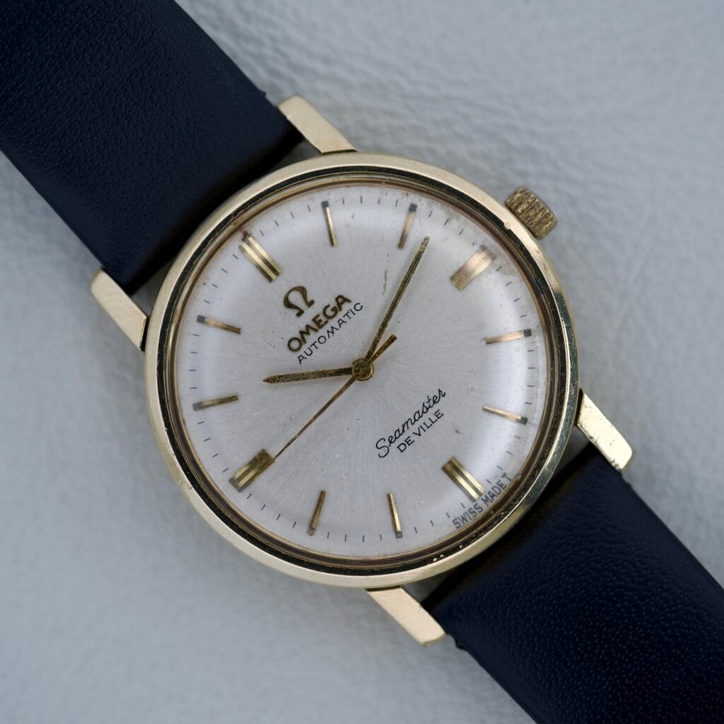 Omega Seamaster Deville Mid-Size wristwatch featuring a 14k gold filled case, sunburst dial, and black leather strap displayed on a white background.