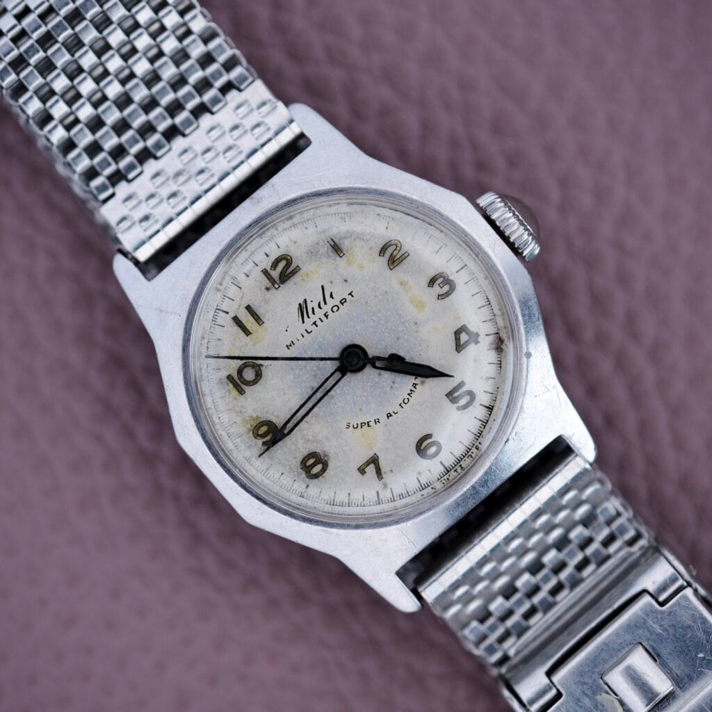 A Mido Multifort Super Automatic Bumper wristwatch with a stainless steel case is shown on a textured mauve surface.