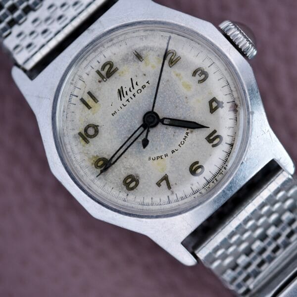 A vintage Mido Multifort Super Automatic Bumper wristwatch featuring a stainless steel case, silver band, and a worn white dial with Arabic numerals.