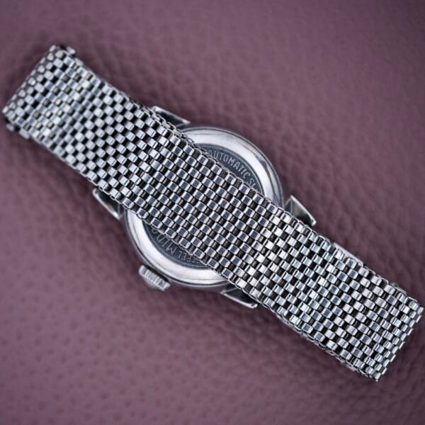 A silver mesh watch band rests on a textured purple surface, with the Mido Multifort Super Automatic Bumper Stainless Steel Case partially visible underneath.