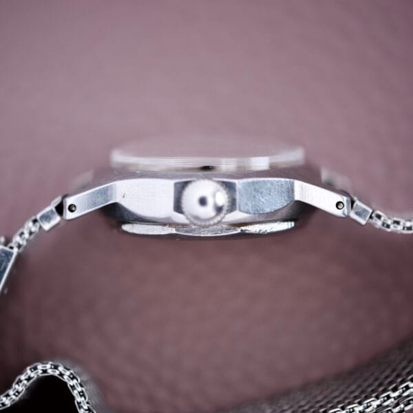 Close-up side view of a Mido Multifort Super Automatic Bumper wristwatch with a stainless steel case, set against a soft pink textured background.