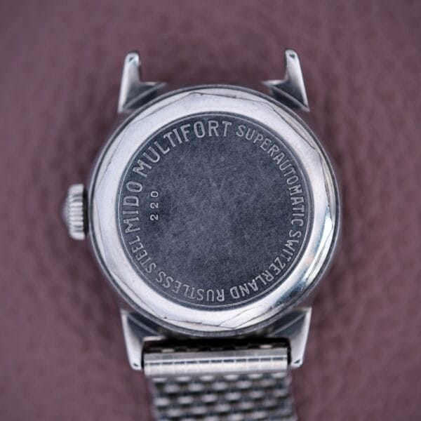 The back of a Mido Multifort Super Automatic Bumper Wristwatch with engraved text and a stainless steel case, resting on a textured mauve surface.
