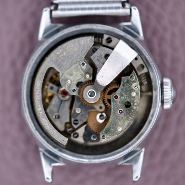 Close-up of an open Mido Multifort Super Automatic Bumper, showcasing its intricate gears and mechanical parts within a polished stainless steel case, highlighting refined craftsmanship on a textured surface.