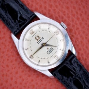 Titus De Luxe 21 Jewels Manual Wind wristwatch with stainless steel case and black leather strap, displayed on an orange textured background.