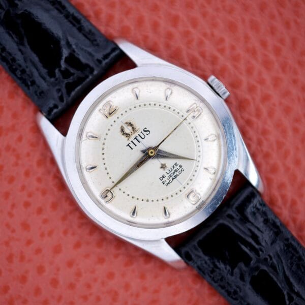 Titus De Luxe 21 Jewels Manual Wind wristwatch with stainless steel case and black leather strap, displayed on an orange textured background.