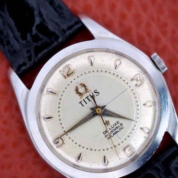 Close-up of a Titus De Luxe 21 Jewels Manual Wind watch with a white dial, black strap, and stainless steel case on a textured red background.