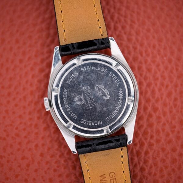 The Titus De Luxe 21 Jewels Manual Wind Stainless Steel Case wristwatch, featuring 21 Jewels engravings on its back, is displayed on a textured orange surface.