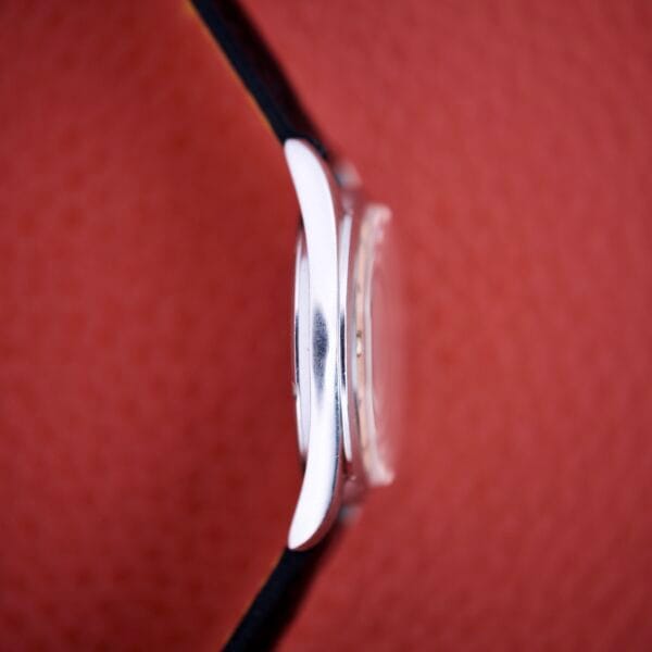 A side view of the Titus De Luxe 21 Jewels Manual Wind wristwatch, featuring a thin stainless steel case against a textured red background.