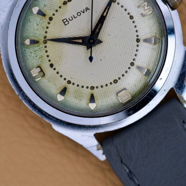 Close-up of a Bulova Waffle Dial Manual Wind Cal. 11ACC US Zone Germany wristwatch featuring a worn cream waffle dial and gray leather strap.