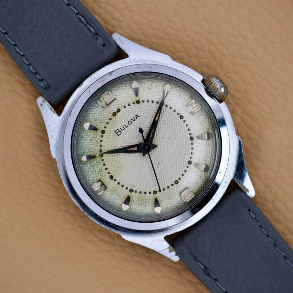 A Bulova Waffle Dial Manual Wind Cal. 11ACC US Zone Germany wristwatch with a cream dial, silver case, and gray leather strap rests on a tan textured surface.