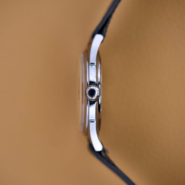 Side view of a Bulova Waffle Dial Manual Wind Cal. 11ACC US Zone Germany wristwatch in silver with a black strap, set against a plain tan background.