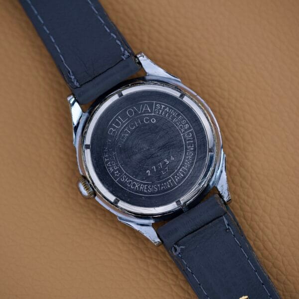 Back of a Bulova Waffle Dial Manual Wind Cal. 11ACC US Zone Germany wristwatch with engraved details, black leather strap, and classic manual wind movement, displayed on a tan surface.