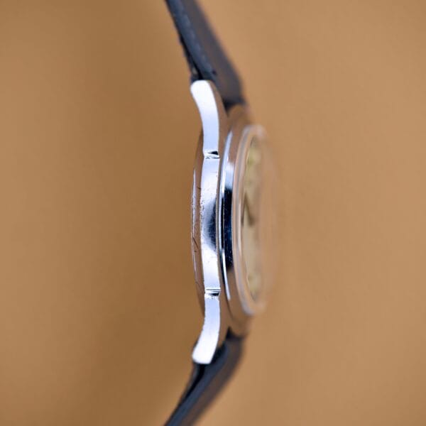 Side view of a silver Bulova Waffle Dial Manual Wind Cal. 11ACC US Zone Germany watch with a black strap on a light brown background.