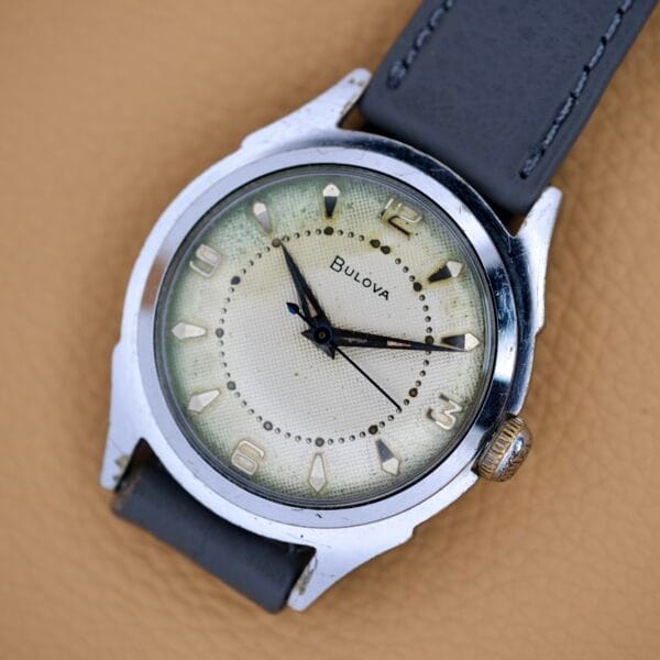 A Bulova Waffle Dial Manual Wind Cal. 11ACC US Zone Germany wristwatch with a gray strap rests on a tan textured surface.