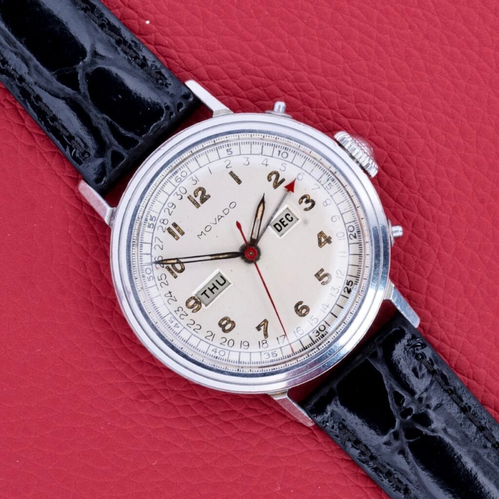 A Movado Triple Calendar Ref. 14819 in stainless steel with manual wind Cal. 475 and black leather strap rests on a red textured surface, elegantly displaying the day and month.