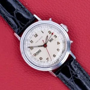 A Movado Triple Calendar Ref. 14819 in stainless steel with manual wind Cal. 475 and black leather strap rests on a red textured surface, elegantly displaying the day and month.