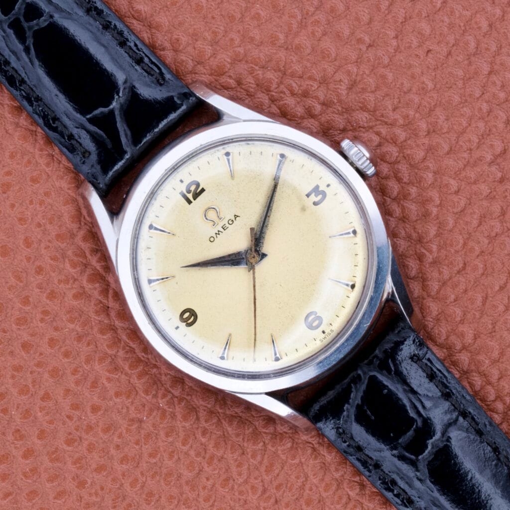 An Omega Ref. 2667-2 in stainless steel with manual wind Cal. 420, cream dial, and black leather strap rests on a textured brown surface.