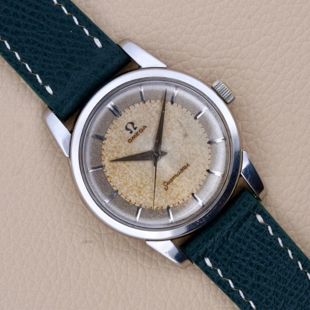 An Omega Seamaster Ref. 2759-7 Manual Wind Cal. 420 in stainless steel with a worn dial and green textured strap rests on a beige surface.