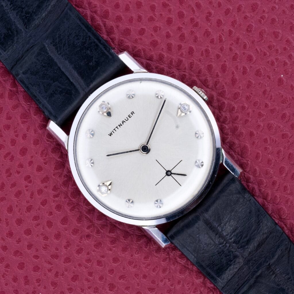 A Wittnauer Diamonds Markers & Sunburst Dial Ref. 1119 Cal. 11BG2 wristwatch with a black leather strap is elegantly displayed on a textured red surface.