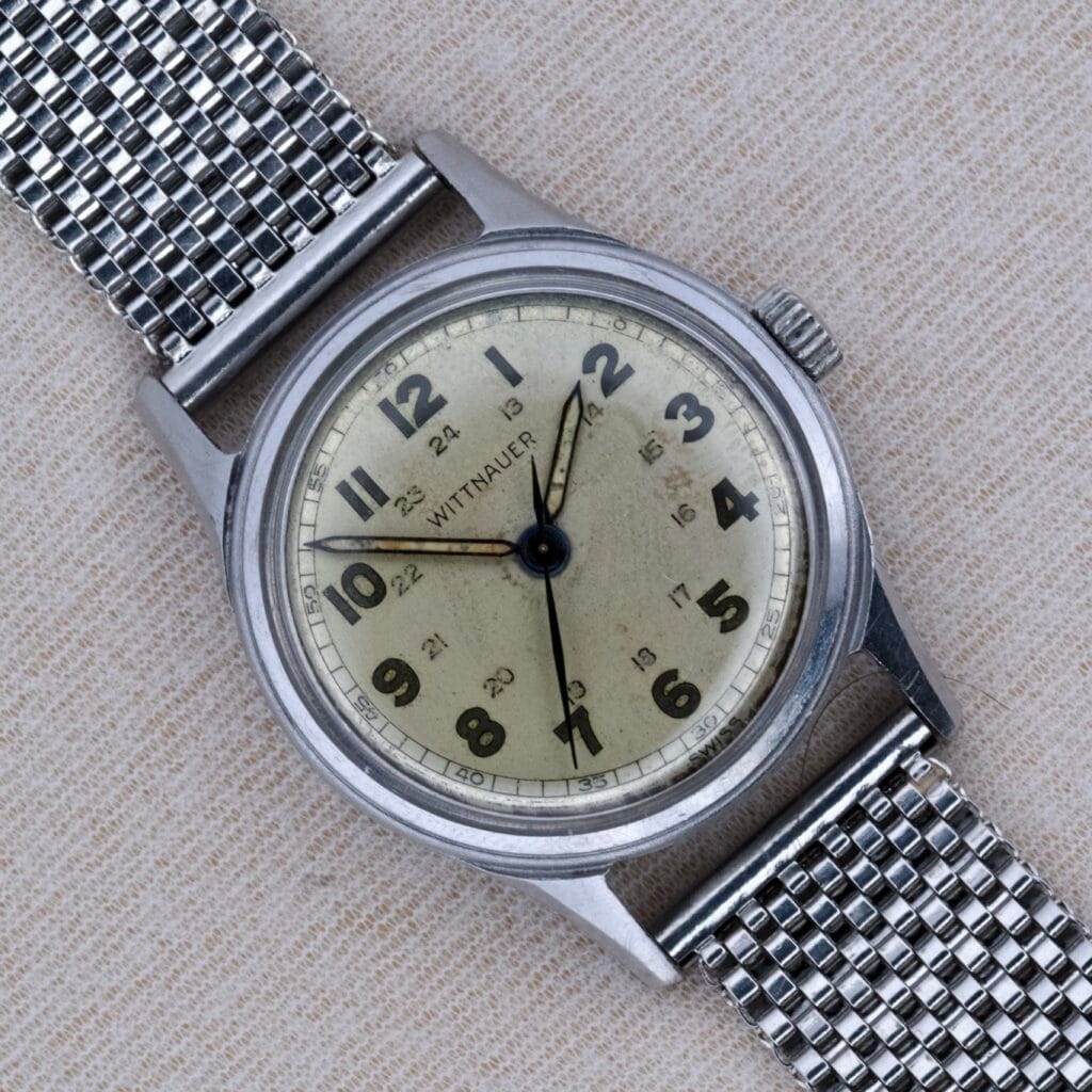 A 1950's Wittnauer Military 24 Hour Dial Manual Wind Cal. 11ES wristwatch with a metal mesh band, showcased on a beige fabric background.