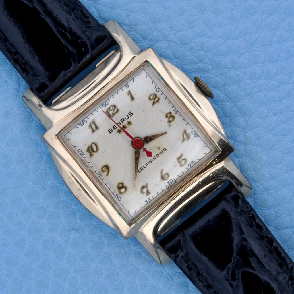 Benrus Three Star Automatic Model CF 1 with a gold plated square case and black leather strap, automatic movement, shown on a light blue textured background.