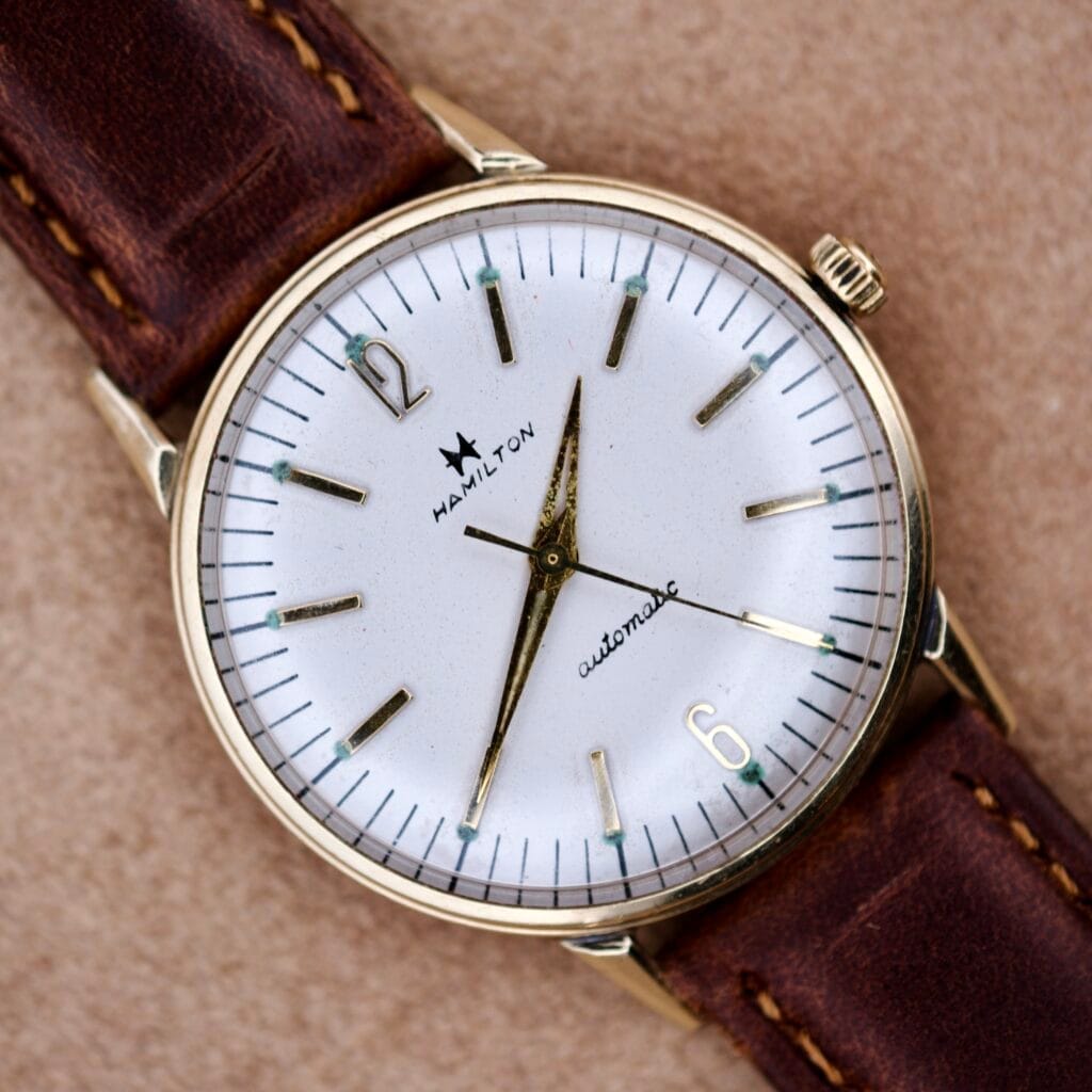 The 1950's Hamilton Automatic features a 10k RGP case and white dial, paired with a brown leather strap, elegantly displayed on a tan surface.