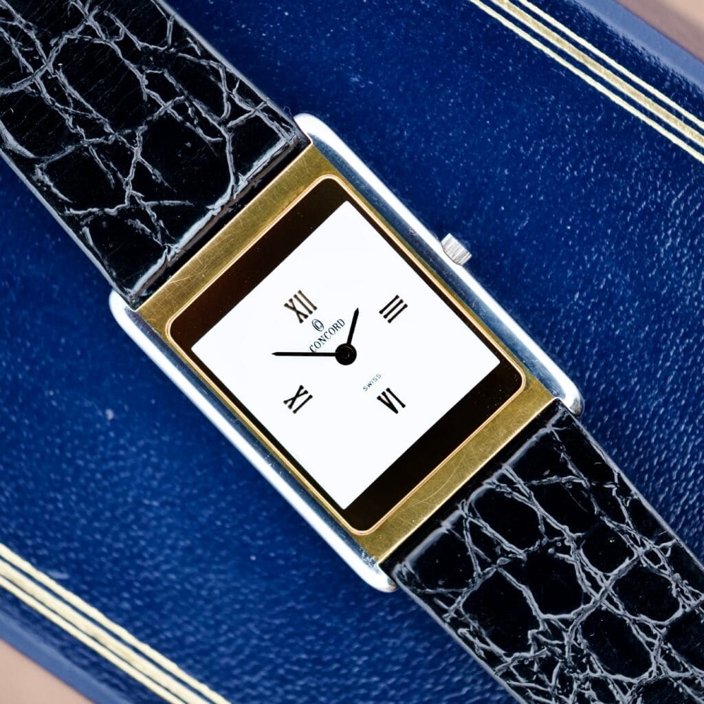 The Concord Delirium Ref. 15-90-667 G Quartz watch features a rectangular case, gold bezel, black leather strap, minimalist Roman numeral dial, and comes with box and papers.