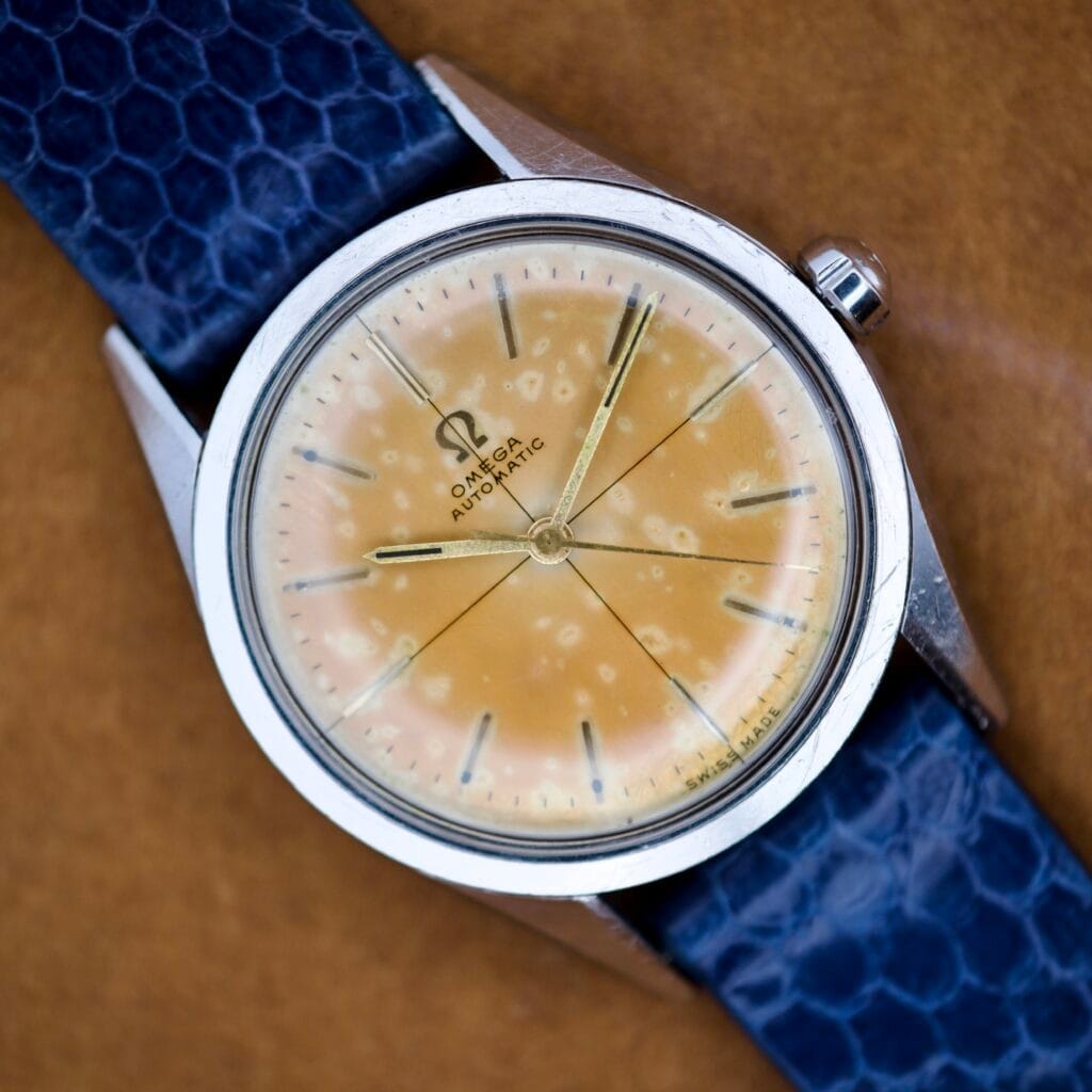 An Omega Automatic Ref. 2802-1 wristwatch featuring a faded, spotty yellow Cross Hair Tropical Dial (Cal. 471) and a textured blue leather strap.