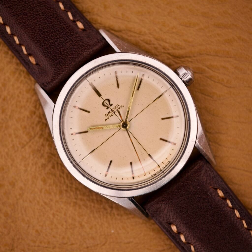 An Omega Automatic Ref. 2802-1 with cross hair cream dial, Cal. 471 movement, and brown leather strap is displayed on a tan surface.