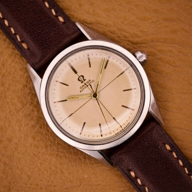 An Omega Automatic Ref. 2802-1 with cross hair cream dial, Cal. 471 movement, and brown leather strap is displayed on a tan surface.