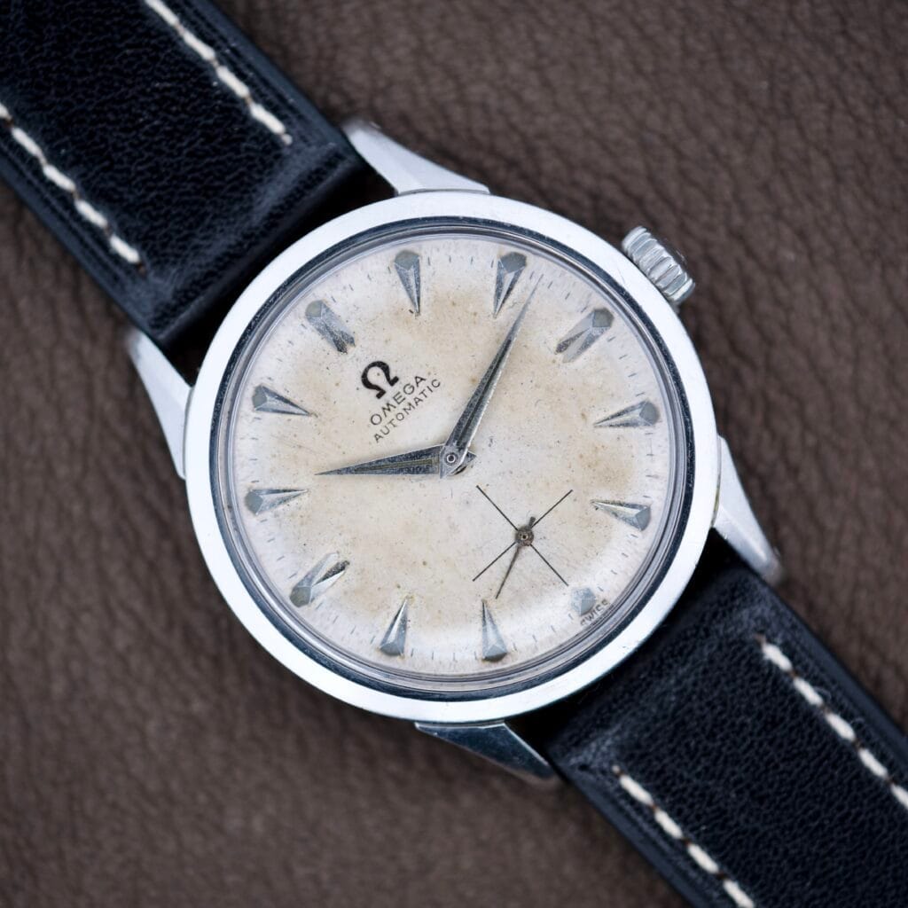Omega Automatic Ref. 6040 with Arctic White Dial, silver markers, and black leather strap displayed on a brown surface. Stainless Steel Cal. 490 model.