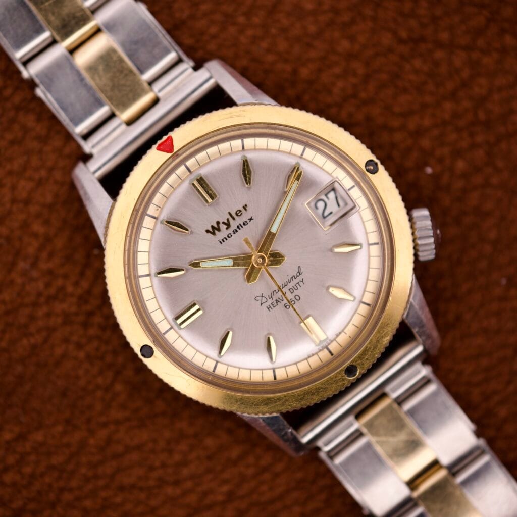 The Wyler Incaflex Dynawind Heavy Duty 660 features a gold bezel, silver dial, date window, and comes with a Riveted Bracelet paired with brown leather for added style.