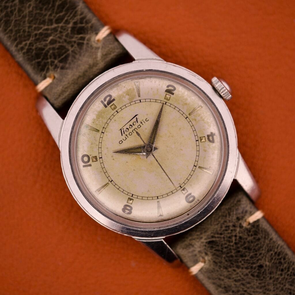 A Tissot Automatic Bumper Ref. 6590 in stainless steel, featuring a worn green strap, rests on a brown leather surface and showcases timeless appeal.
