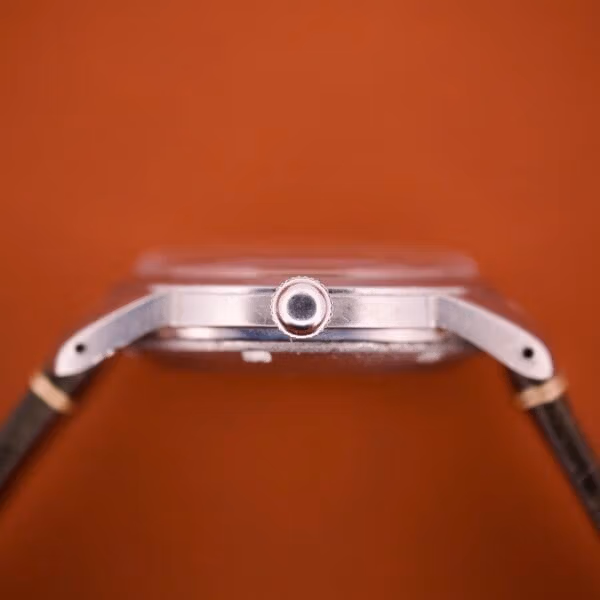 Crown profile of the Tissot 6590 showing the signed grooved steel crown and the slim stainless midcase against an orange backdrop.