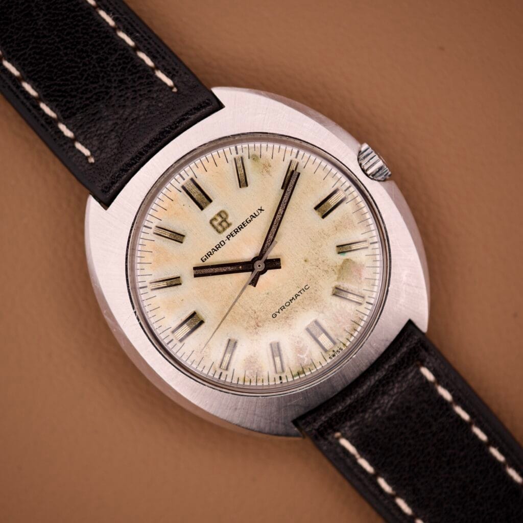 The Girard Perregaux Gyromatic Stainless Steel Cushion Case Ref. 9382 GA features a worn beige dial, black leather strap, and rests on a brown surface, with its stainless steel case adding classic charm.