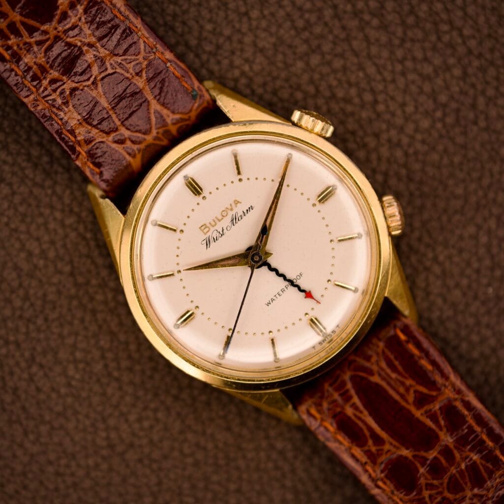 Bulova Wrist Alarm with gold tone case and manual wind Cal. 11AERC movement, paired with a brown leather strap, displayed on a textured brown surface.