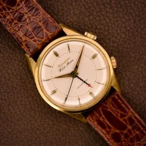 Bulova Wrist Alarm with gold tone case and manual wind Cal. 11AERC movement, paired with a brown leather strap, displayed on a textured brown surface.