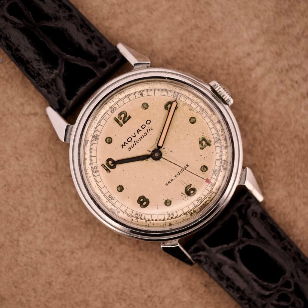 Movado Automatic Bumper FAB. Suisse Ref. 16151 Stainless Steel watch featuring a cream dial with black numerals and a black leather strap, displayed on a tan background.