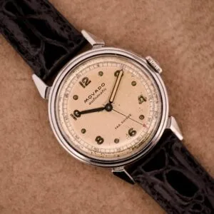 Movado Automatic Bumper FAB. Suisse Ref. 16151 Stainless Steel watch featuring a cream dial with black numerals and a black leather strap, displayed on a tan background.