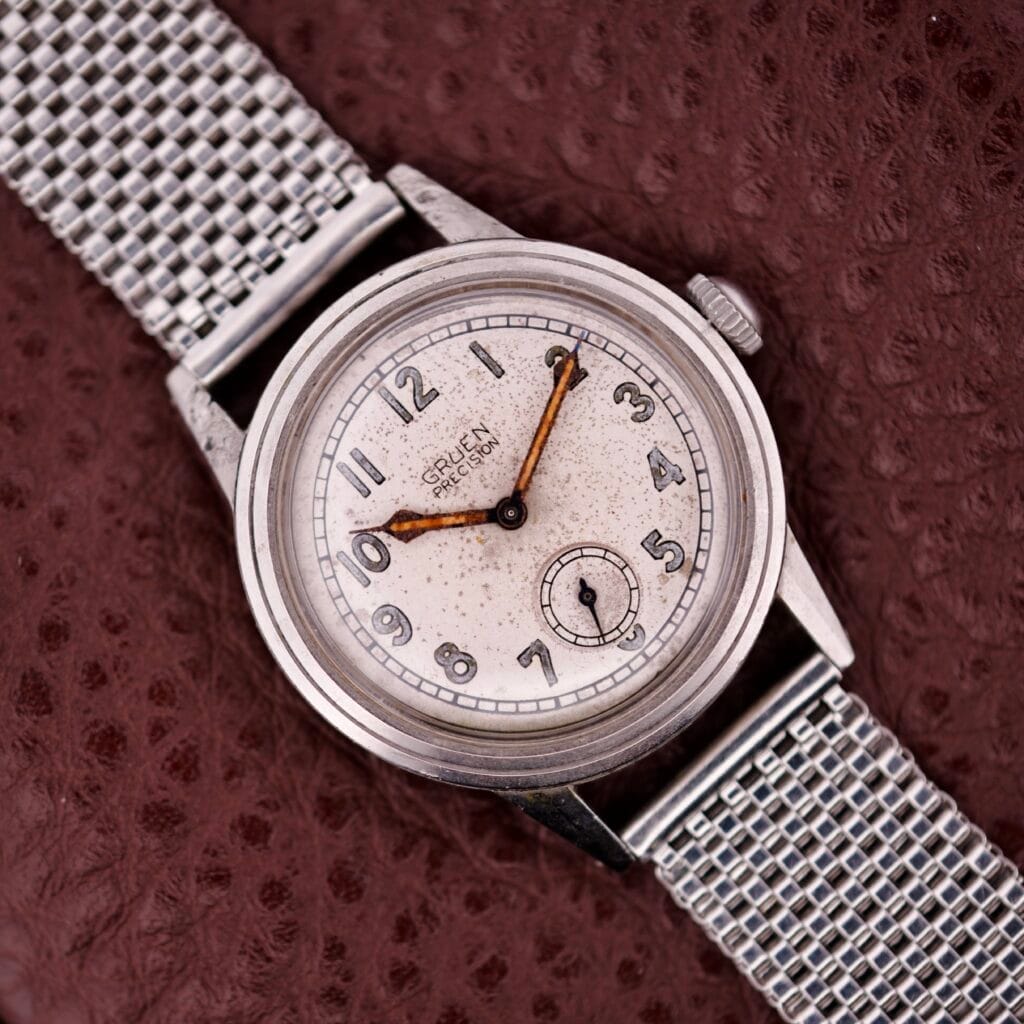 A vintage Gruen Precision Auto-Wind with Military Dial in a stainless steel case, shown on a textured brown surface.