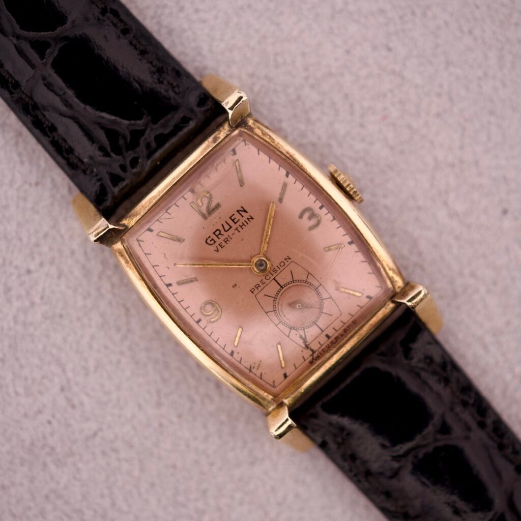 Gruen Veri-Thin Precision Art Deco Lugs & Salmon Dial Ref. 430-457 gold wristwatch with a rectangular salmon dial and black leather strap, elegantly displayed on a light surface.