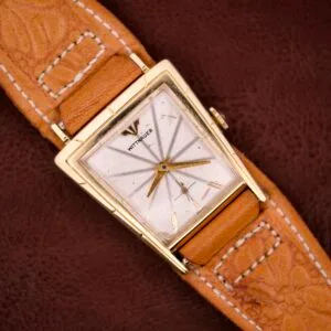 A vintage Wittnauer Asymmetrical Case Ref. 2025 watch with a gold case, tan leather strap, and manual wind Cal. 9E movement is displayed on a brown textured background.