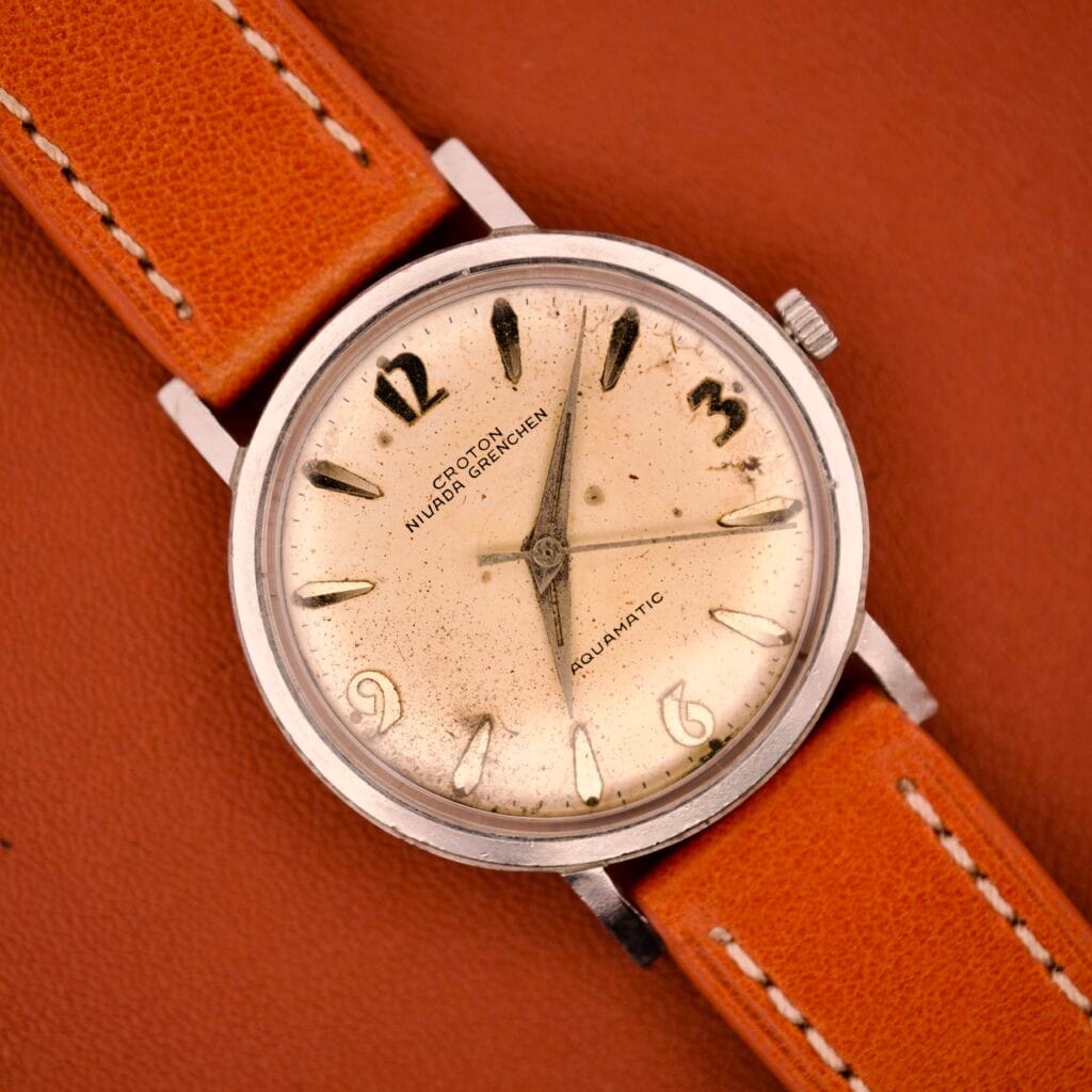 The Croton Nivada Gretchen Aquamatic Stainless Steel ETA Cal. 2451 watch, with a tan leather strap, is displayed elegantly on a brown surface.