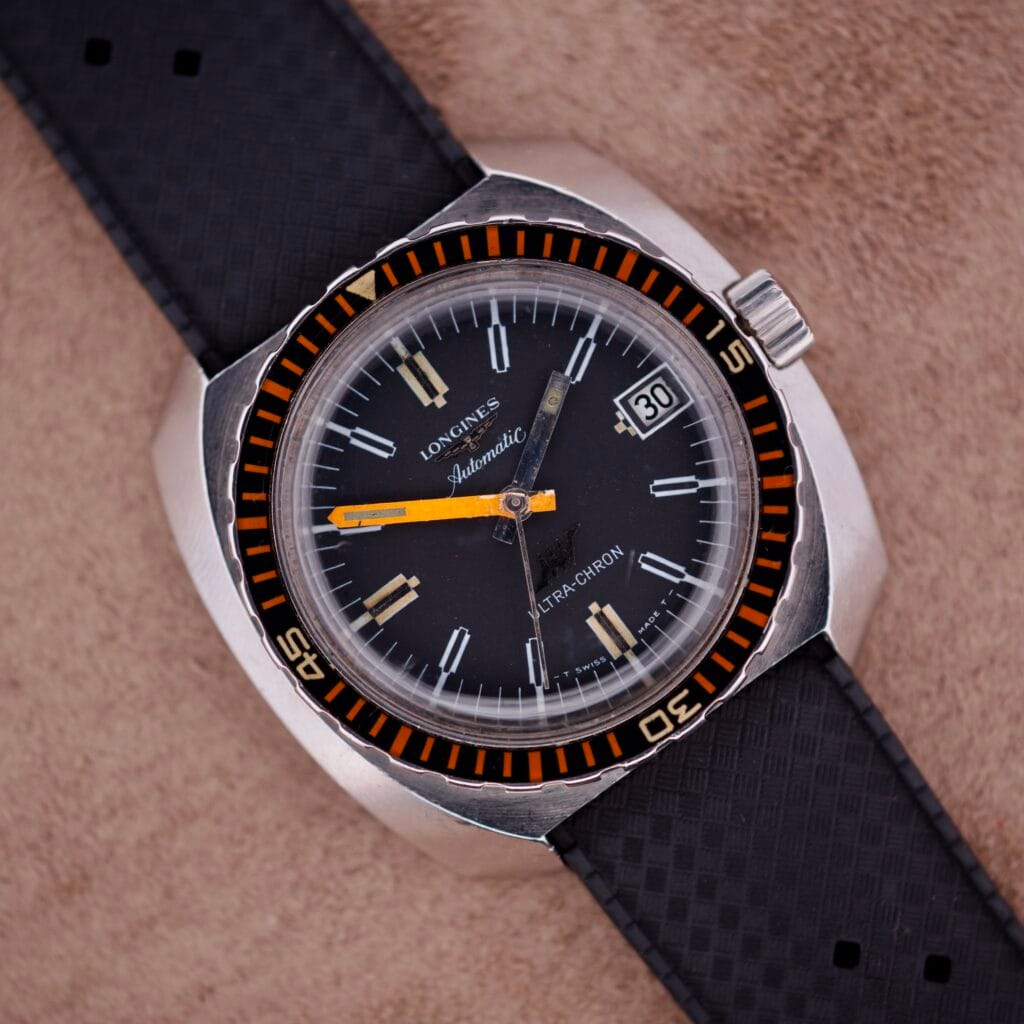 Vintage Longines Ultra Chron Diver Ref. 7970-4 with black dial, orange accents, and a black textured strap on a tan surface, featuring the iconic Balkelite bezel in stainless steel.