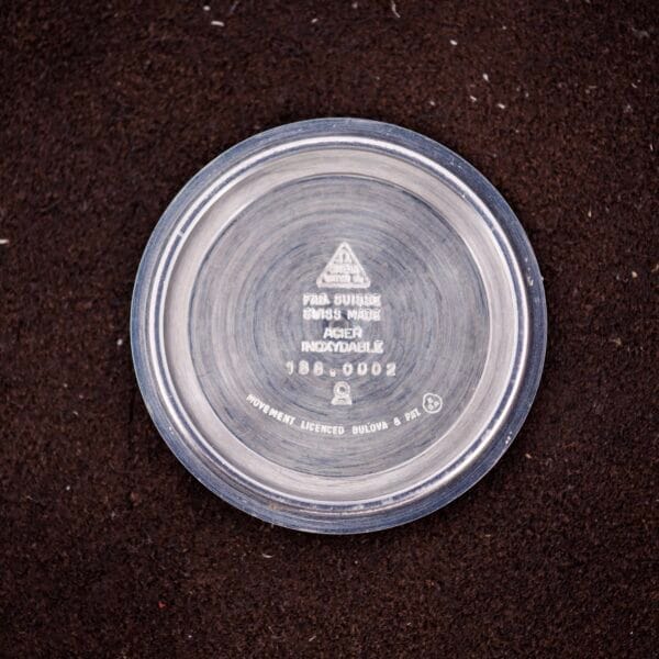 A round metal watch case back with engravings, likely from an Omega Seamaster Speedsonic F300 Ref. 188.0002 Chronograph Original Bracelet Spanish Date, rests on a dark textured surface.